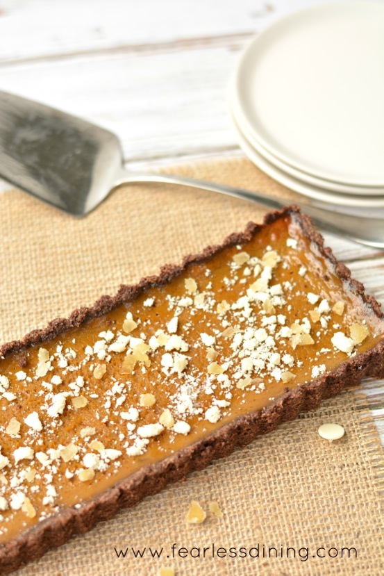 Gluten free pumpkin tart with white chocolate, candied ginger, and dark rum. The tart is on a table with a serving utensil and plates next to it.