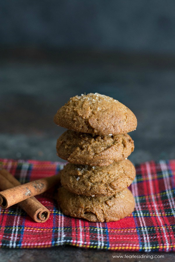 Four gluten free ginger cookies stacked on top of each other on a red plaid napkin. Two cinnamon sticks are next to the stack.