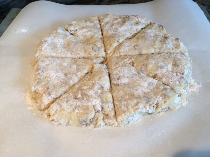 A round of scone dough on parchment paper. The dough is cut into wedges.