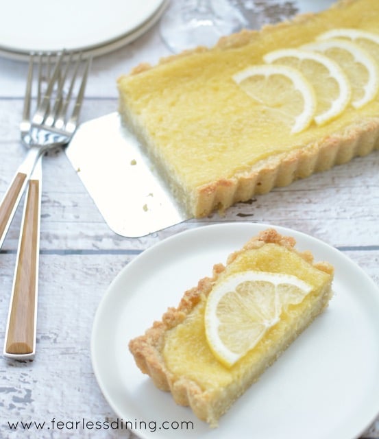 Gluten Free Lemon Tart on a white table. A slice of tart is on a plate with forks next to it.
