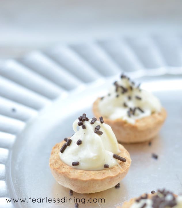 The close up of two Gluten Free Baked Cannoli Bites with chocolate sprinkles on them. The cannoli cups are on a silver platter.