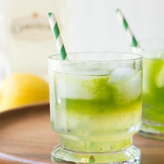 2 cocktail glasses of tequila lemonade with green striped straws sit on a wood serving platter.