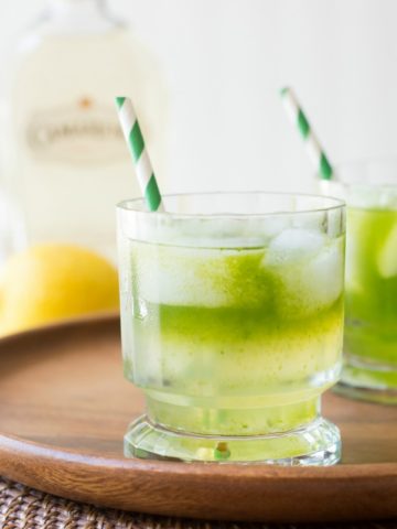 2 cocktail glasses of tequila lemonade with green striped straws sit on a wood serving platter