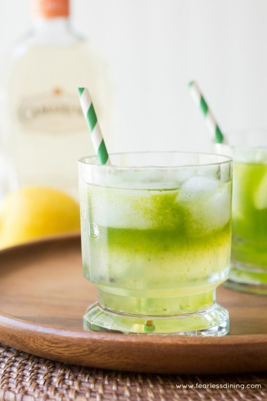 2 cocktail glasses of tequila lemonade with green striped straws sit on a wood serving platter