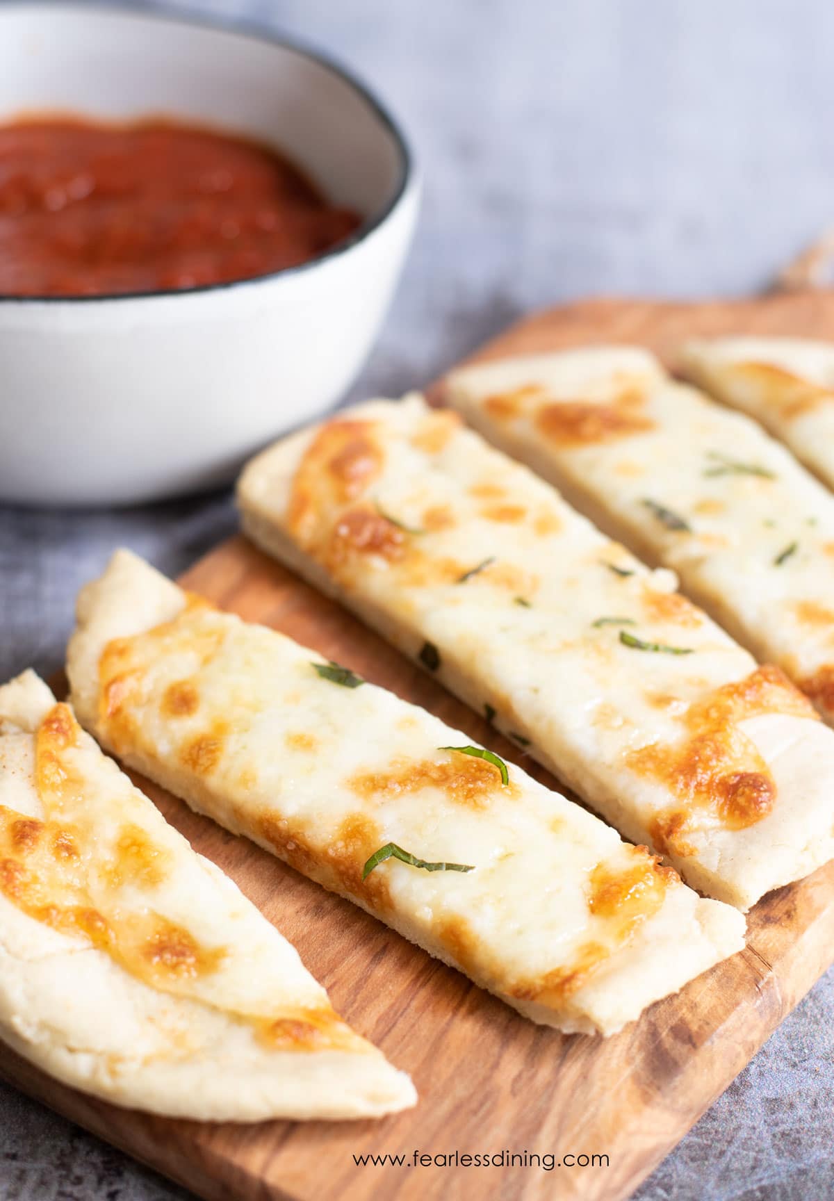 A cheese topped sliced gluten free flatbread on a wooden cutting board.