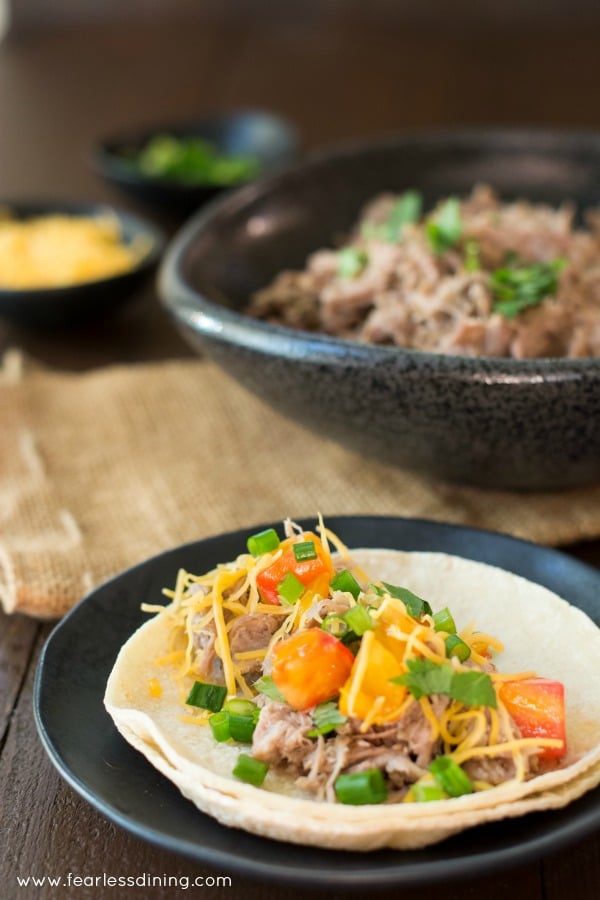 Soft pulled pork taco on a plate with a bowl of pulled pork in the background.