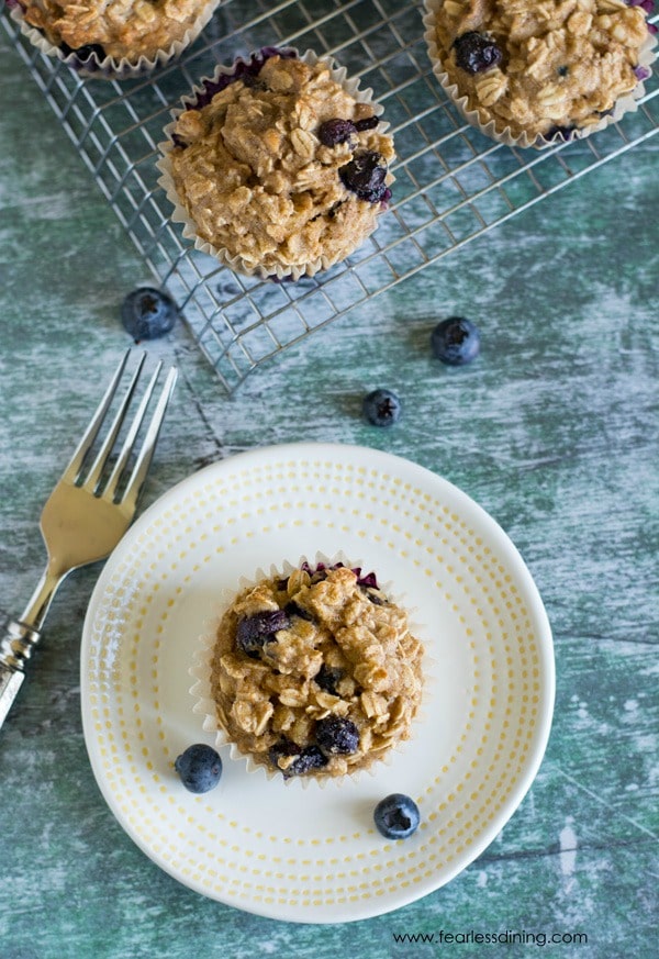 Top view of a blueberry oat muffin on a white plate. The rack of muffins is next to the plate.