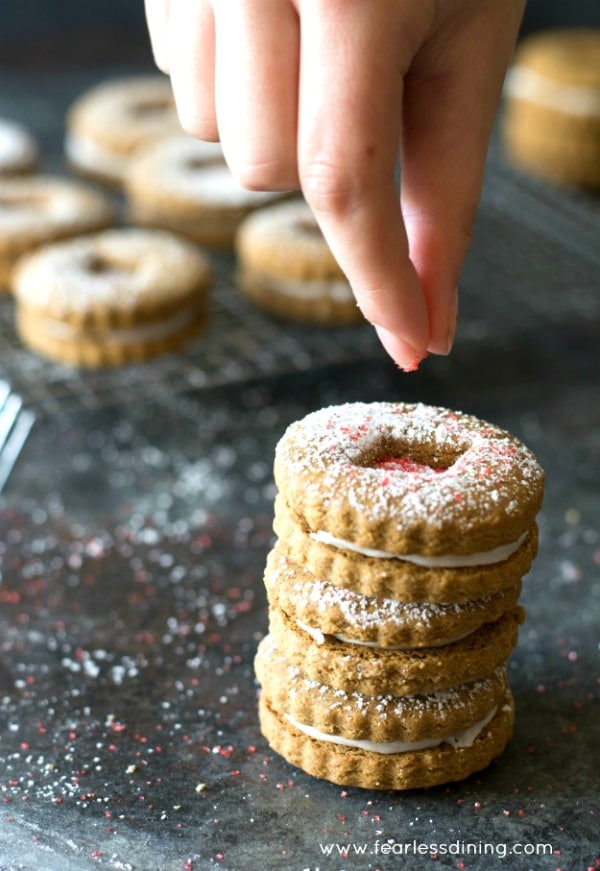Sprinkling on red sprinkles onto gluten free gingerbread linzer cookies in a stack