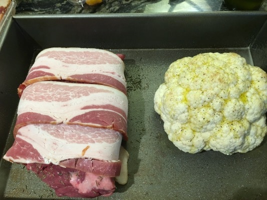 Roasting a Whole Cauliflower Head next to a roast wrapped in bacon in a pan.