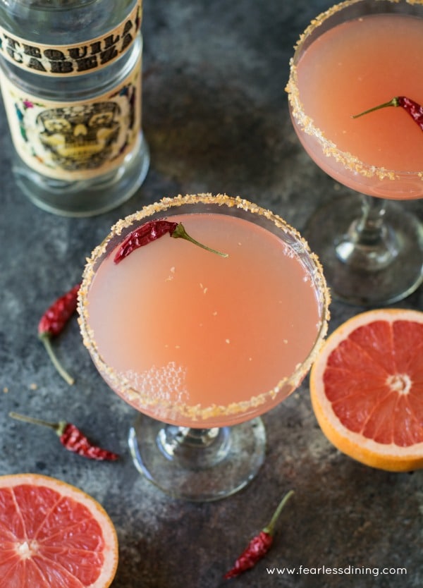 Pink Grapefruit Margaritas with Sriracha Salt are perfect for a party! You are going to love the unique sweet and spicy combination of flavors of the grapefruit and sriracha salt in this refreshing pink grapefruit margarita recipe. Two Pink Grapefruit Margaritas with Sriracha Salt ready to drink. A bottle of tequila is in the background.