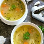 Two bowls of chicken soup next to carrots and a garlic bulb.