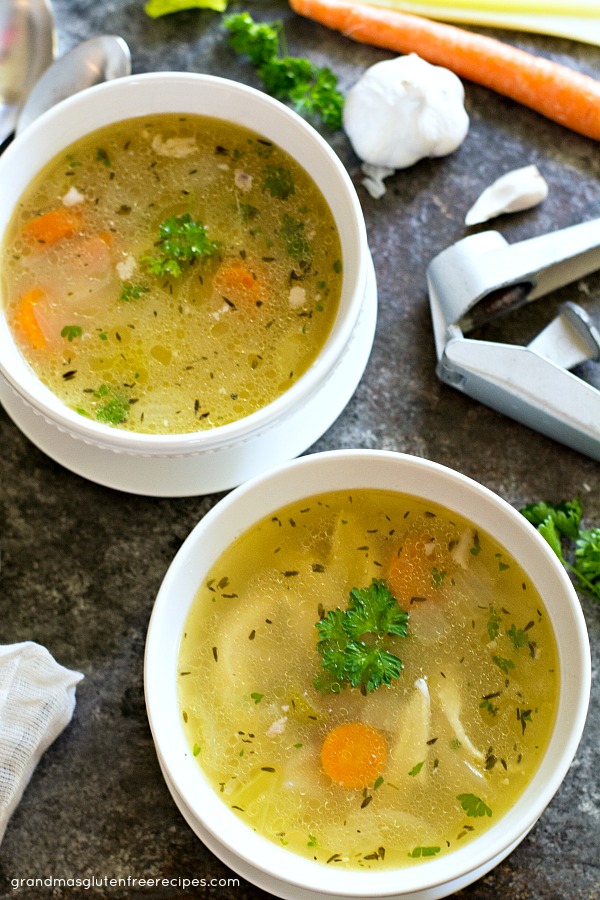 Two bowls of chicken soup next to carrots and a garlic bulb.