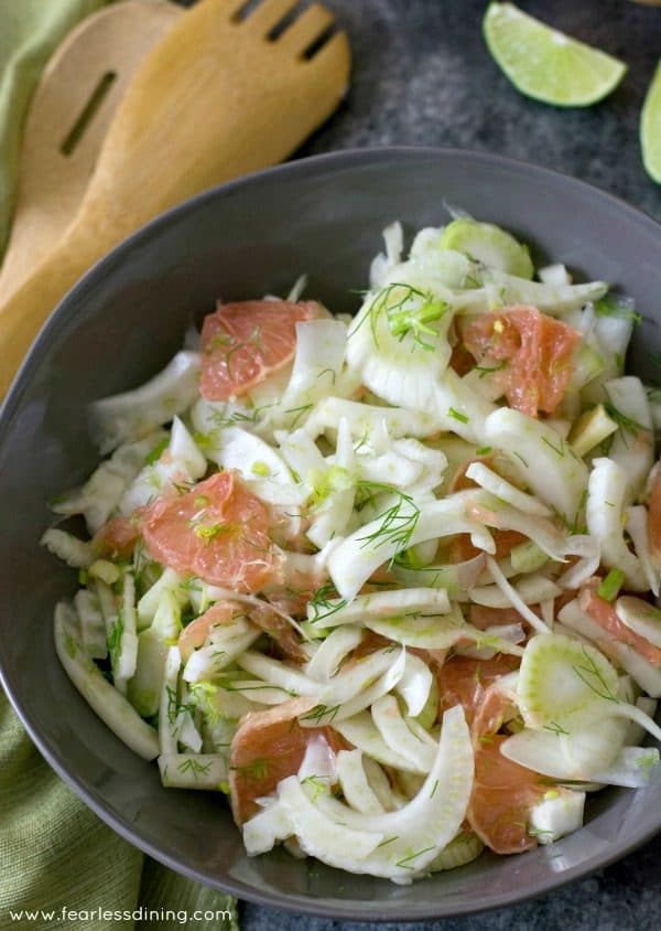 Top view of a large bowl of pink grapefruit and fennel salad. Serving spoons and lime wedges are along the side.