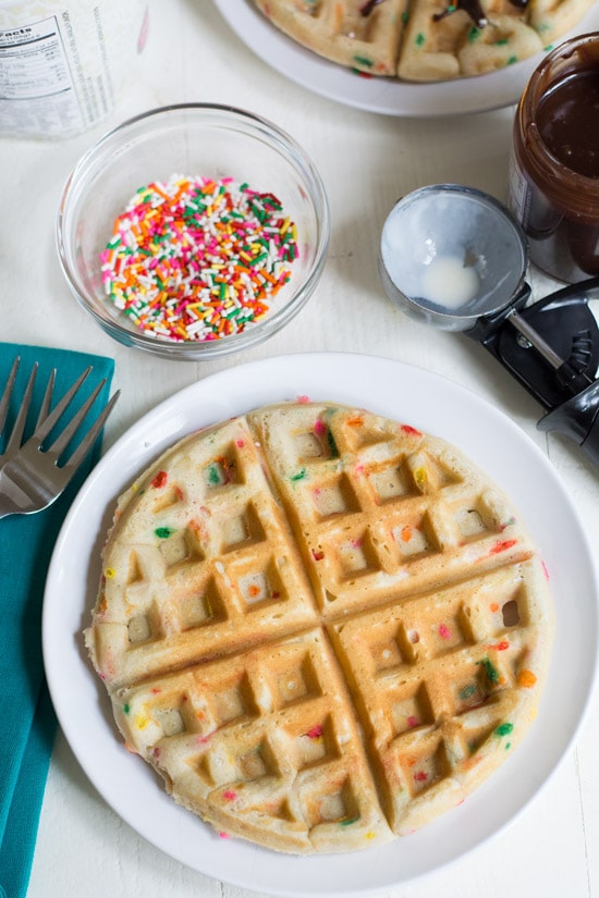 Top view of a cake batter waffle on a plate with a bowl of sprinkles, an ice cream scoop and hot fudge next to the plate.