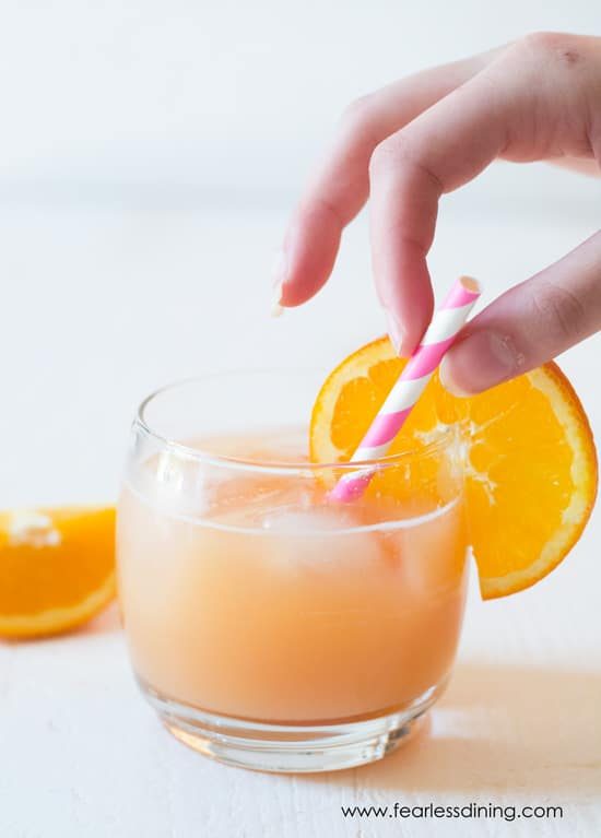 Juice in a glass with an orange garnish. A hand is grabbing the pink and white striped straw.