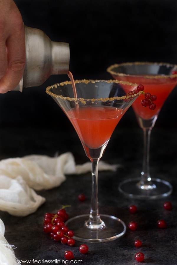 A shaker is pouring the red currant margarita into a tall martini glass.