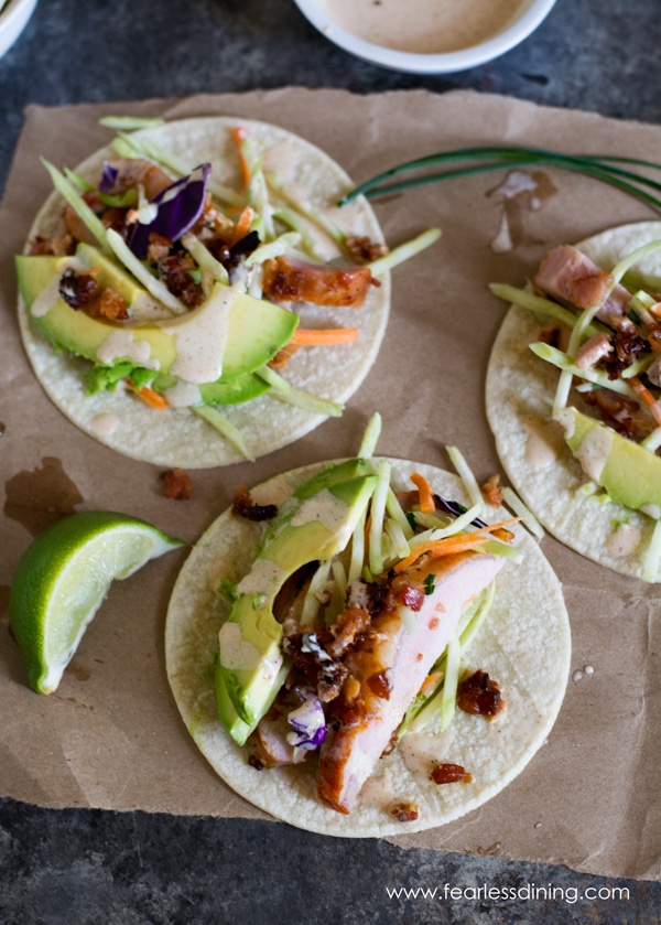 Looking down at three tacos. The tacos are on a brown paper bag. They are topped with avocado and a slice of lime.