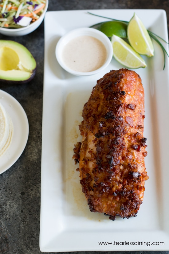 A rectangular white plate with a grilled pork tenderloin on it. Cut limes and a dipping sauce are also on the plate.