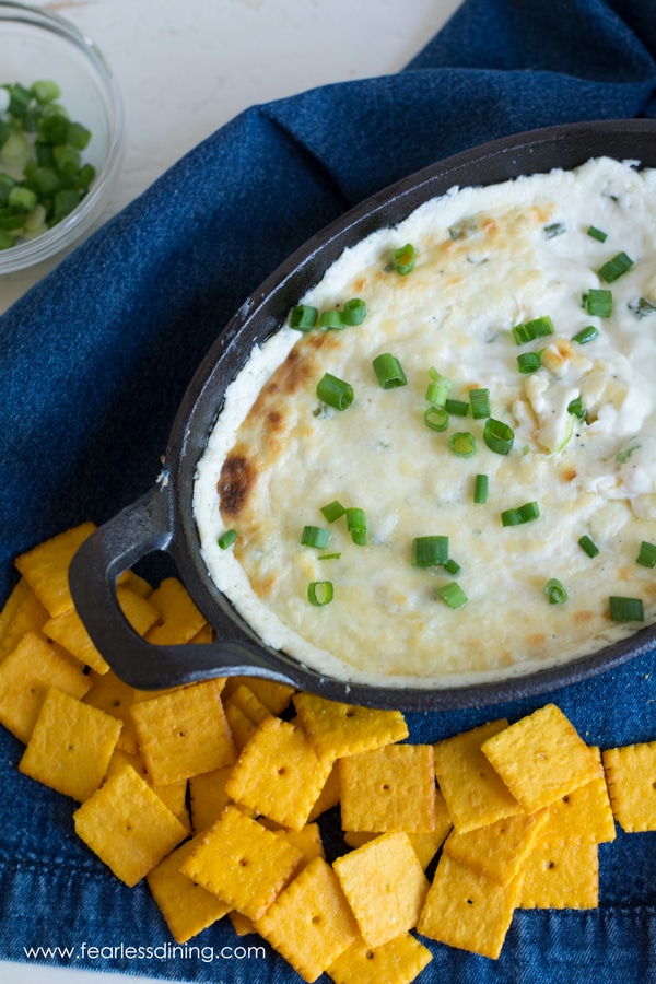 A cast iron baking dish filled with a melted cheese dip. The dip is topped with scallions. Cheese crackers are next to the dish.