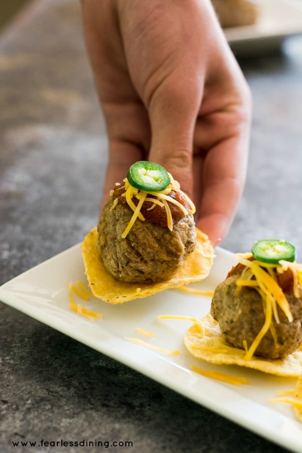 A hand grabbing a tortilla chip with a meatball on top. The meatball is topped with salsa, cheese and a jalapeño slice.