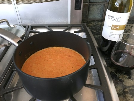 Red pepper soup cooking on the stove in a pot. A bottle of wine is next to the stove