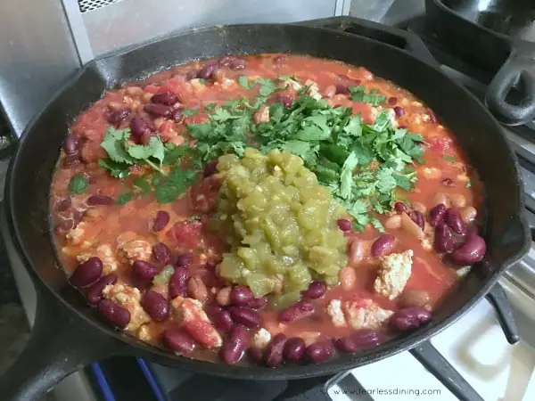 chili cooking in a skillet. Green chilis and cilantro are on top being cooked in.