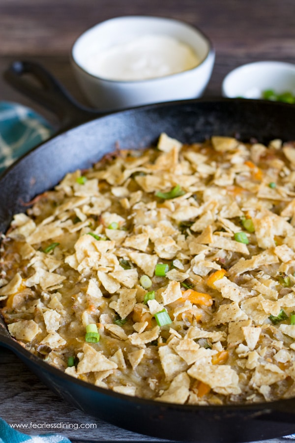 a cast iron skillet full of cheesy funeral potatoes. Sour cream and scallions are in small bowls behind the skillet.