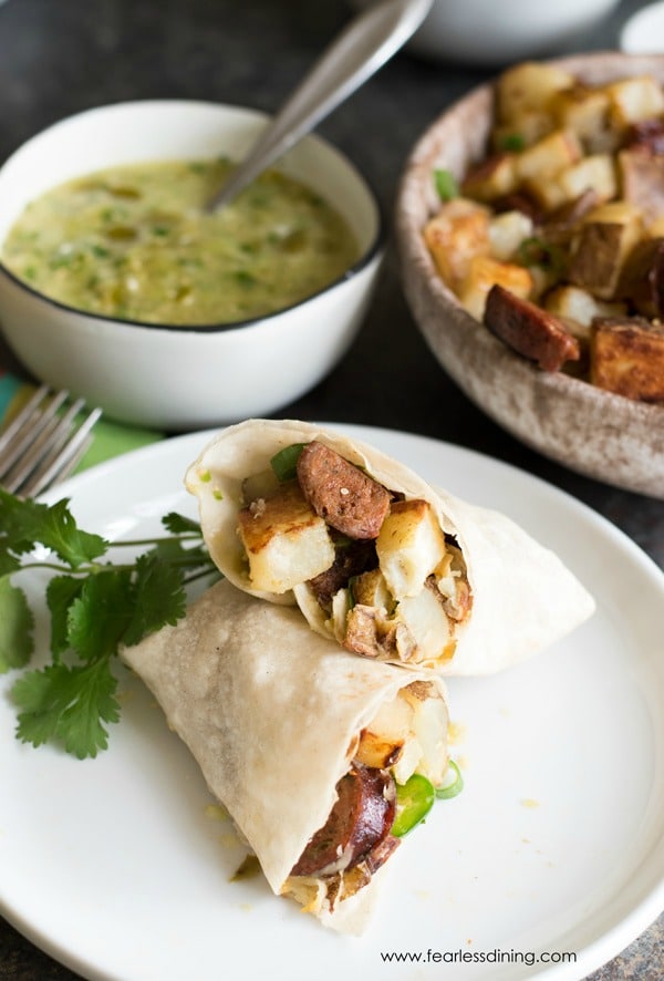 A potato chorizo breakfast burrito cut open. Bowls with potatoes and salsa verde are behind the plate