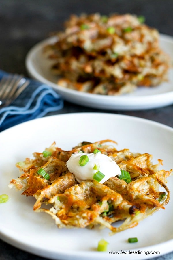 A crispy hash brown waffle on a white plate. A stack of hash brown waffles are on a plate behind this.