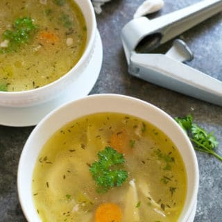 Two bowls of chicken soup with a garlic press and garlic cloves next to the bowls.