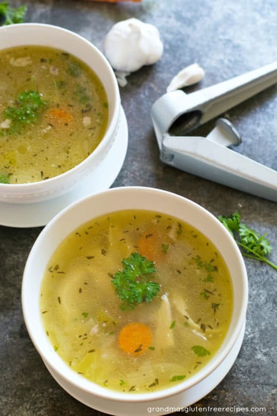 Two bowls of chicken soup with a garlic press and garlic cloves next to the bowls.