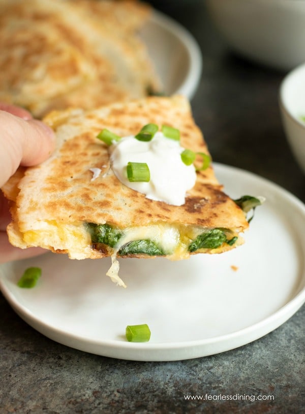 A hand holding a slice of quesadilla over a white plate.