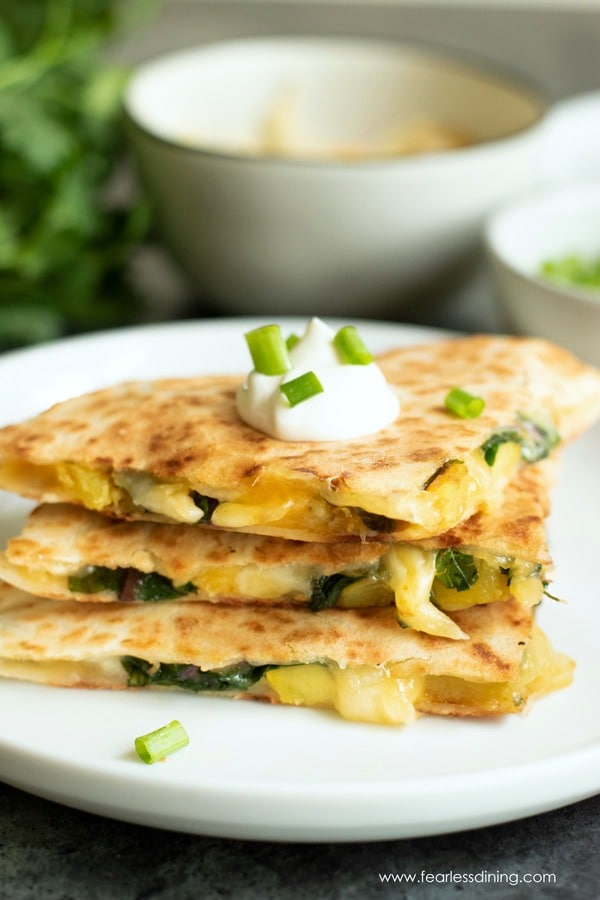 A stack of cut delicata squash quesadillas on a white plate. The quesadillas are garnished with sour cream and scallions.