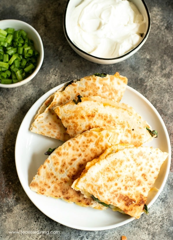 Sliced pieces of quesadilla on a plate. A bowl of sour cream and a bowl of scallions are next to the plate.