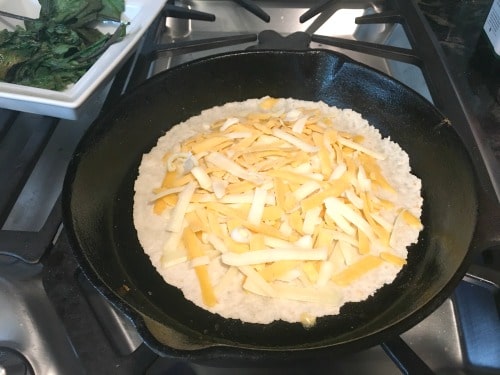 A tortilla in a cast iron pan cooking with shredded cheese on top.