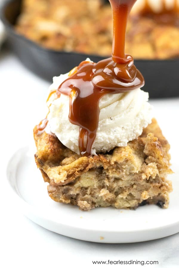 A slice of gluten free banana bread pudding on a plate. It is topped with a scoop of ice cream and hot caramel is being drizzled on top.