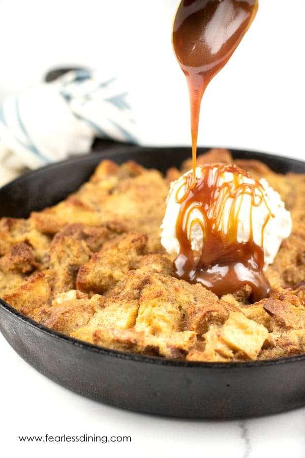 A spoon drizzling caramel over the skillet of gluten free banana bread pudding.