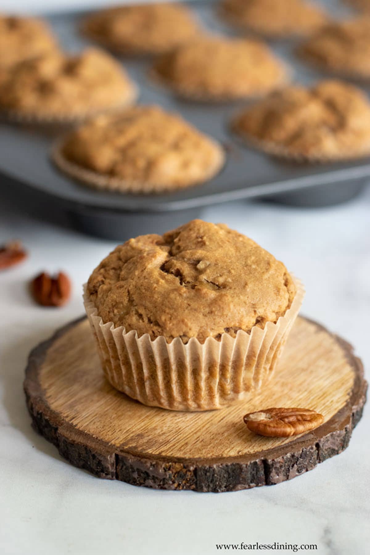 A gluten free banana muffin on a wooden coaster next to a tray of muffins.