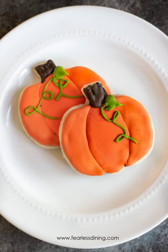Two gluten free pumpkin shaped cookies decorated with royal icing on a plate.