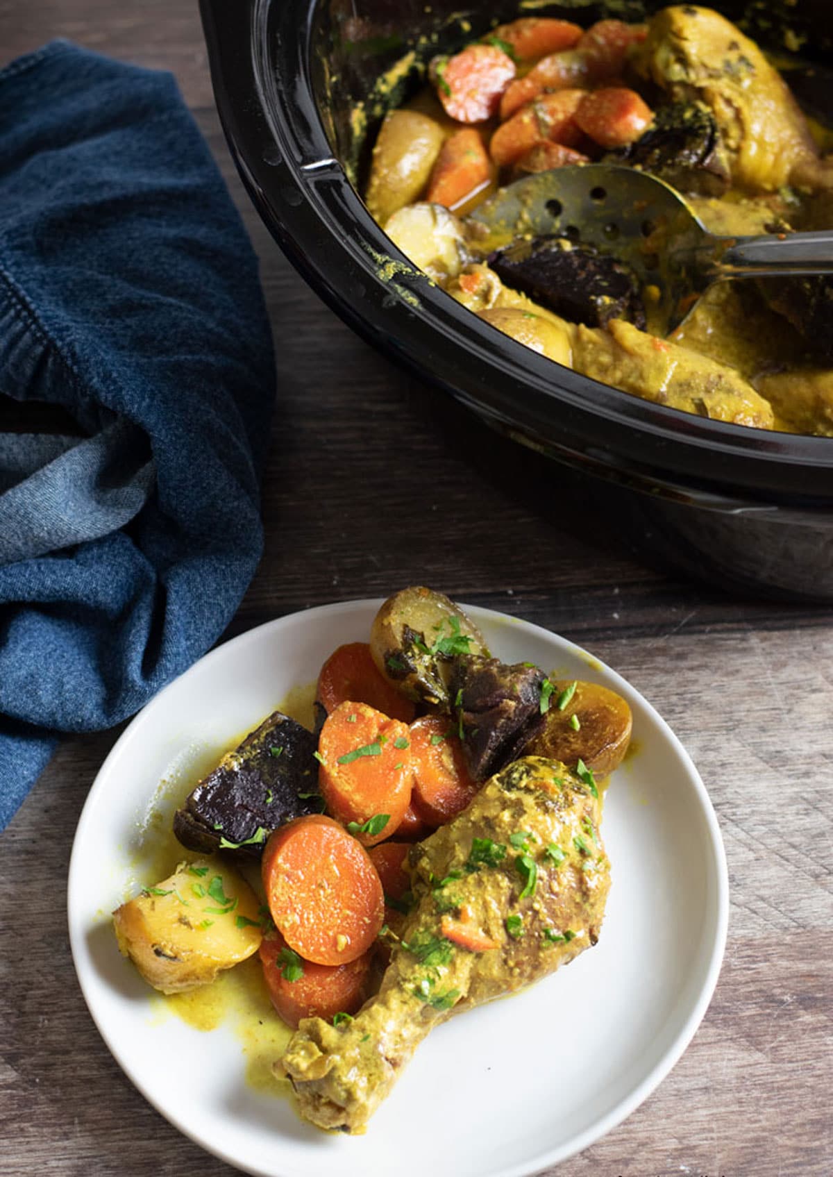 Top view of a plate with a curry chicken leg and root vegetables next to the slow cooker.