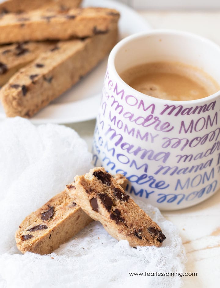 Gluten free chocolate chip biscotti next to a coffee mug. A plate of biscotti is also behind the mug.