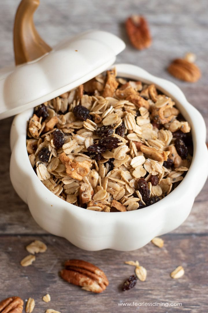 Apple granola in a white ceramic pumpkin bowl. The lid is laid slanted next to the bowl.