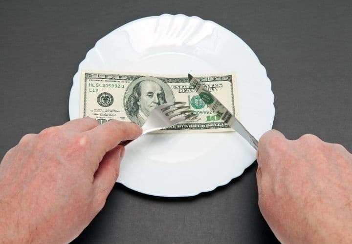 A paper 100 dollar bill on a plate being cut with a fork and knife.