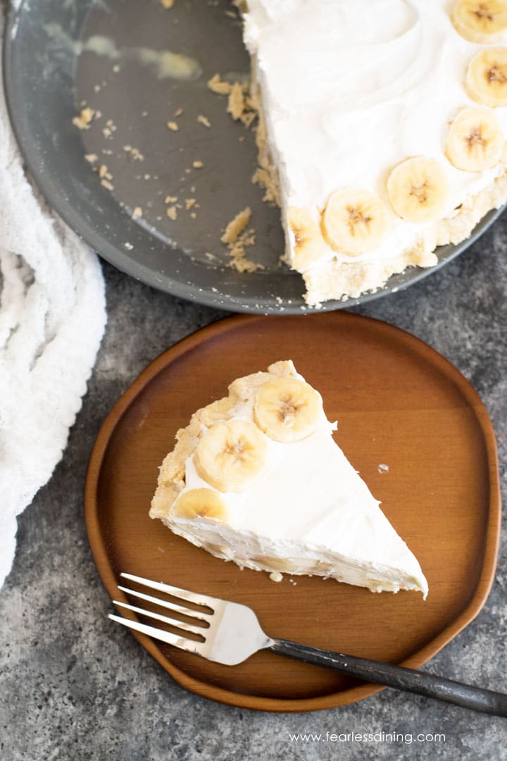 The top view of a slice of banana cream pie on a wooden plate next to the pie tin of pie.