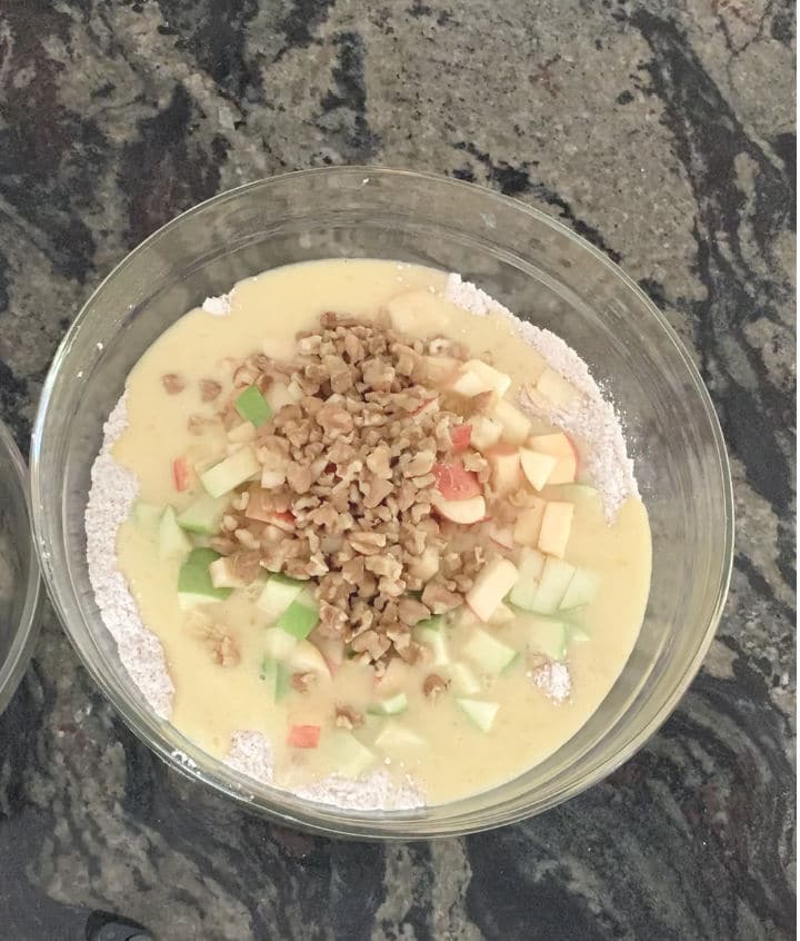 A bowl with dry ingredients, wet ingredients on top, and the apples and walnuts. It is ready to be mixed.