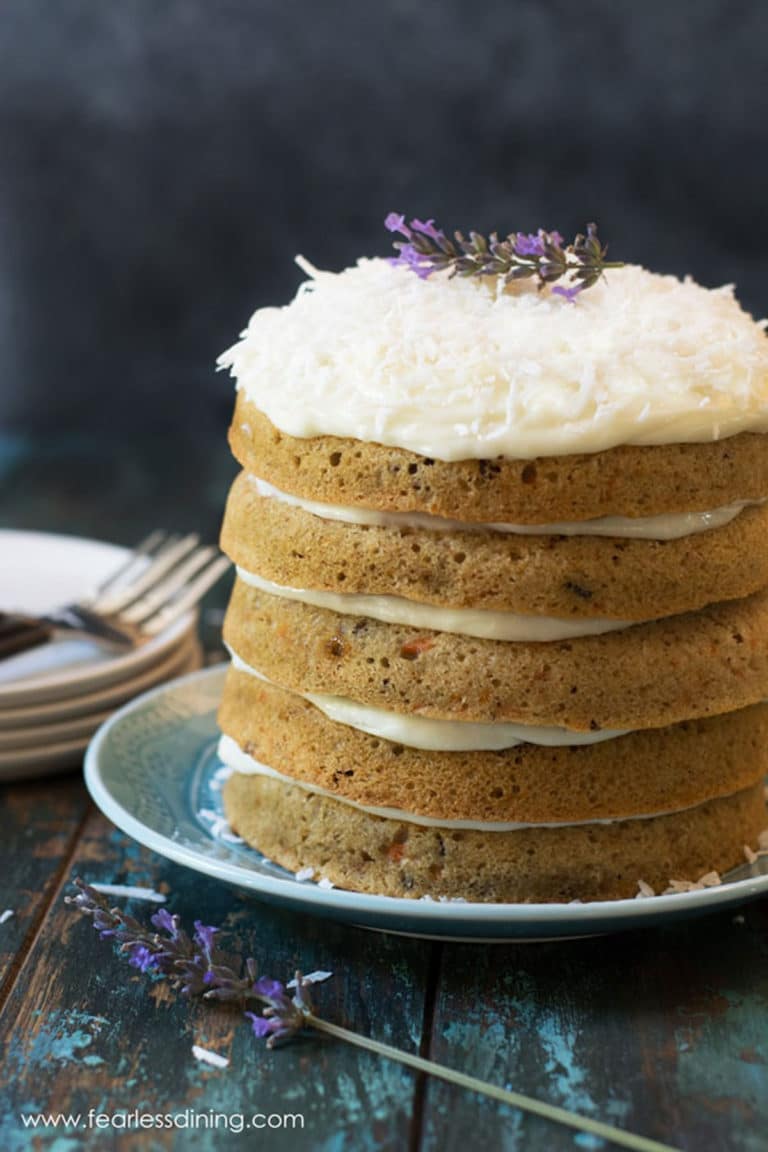 A fancy carrot cake with cream cheese frosting. The cake is topped with shredded coconut and a lavendar flower.