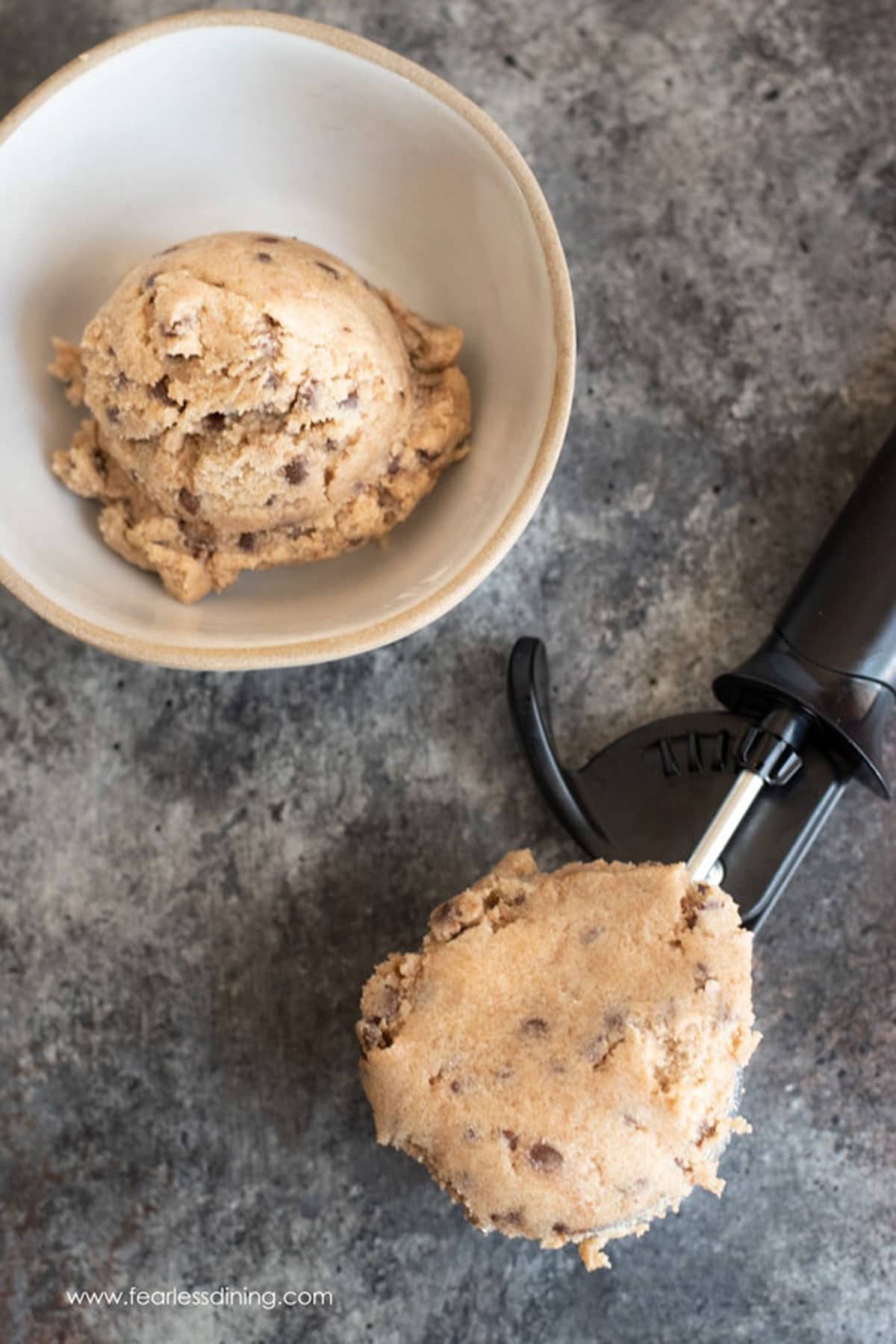 The top view of a bowl of cookie dough next to a scoop.