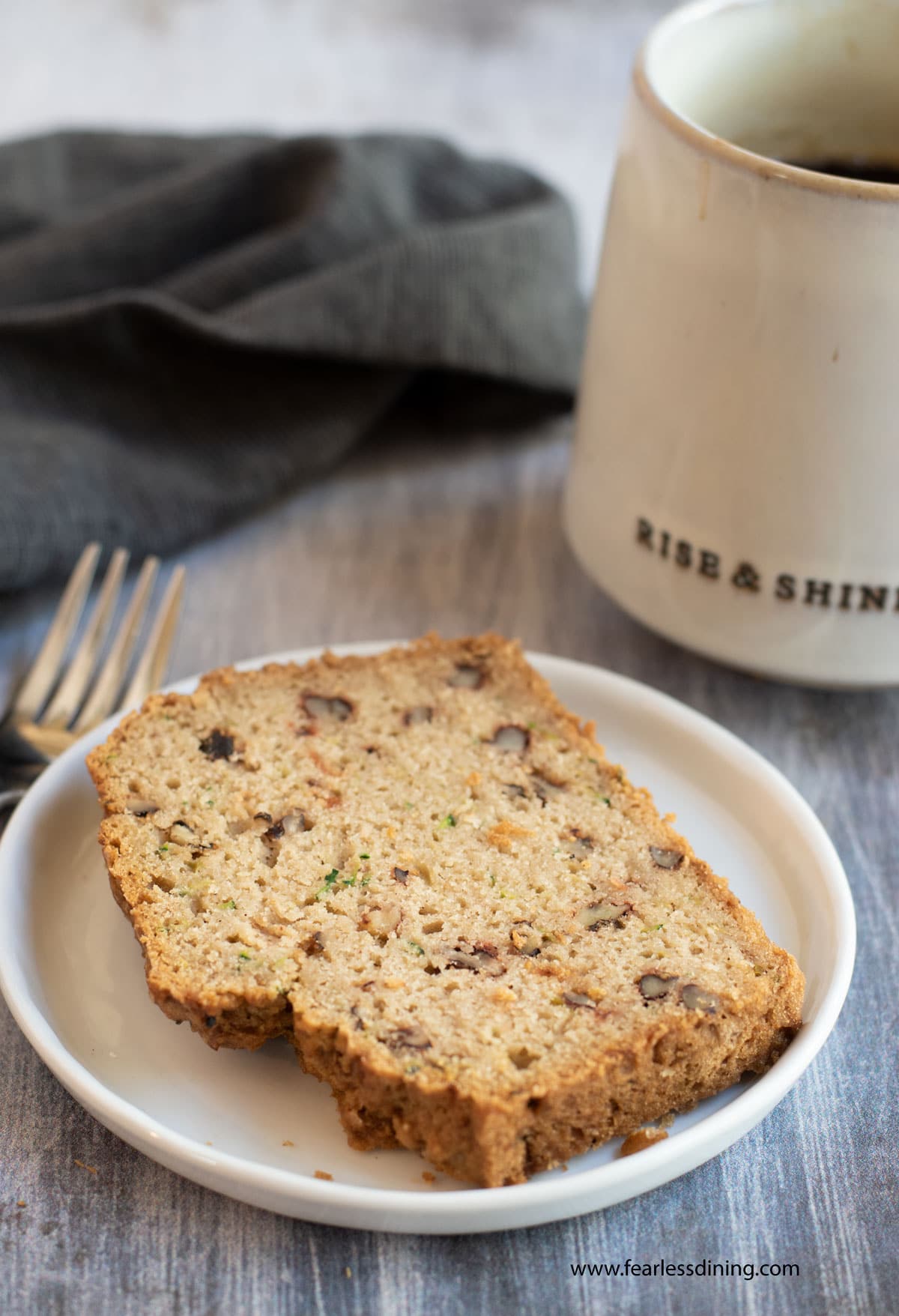 A slice of zucchini bread on a plate next to a mug of coffee.