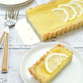 A slice of lemon tart on a white plate next to the whole tart.
