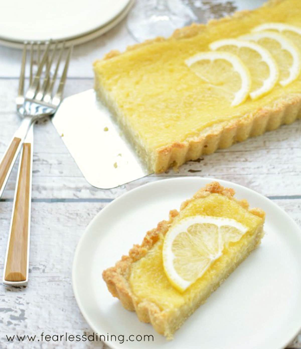 A slice of lemon tart on a white plate next to the whole tart.